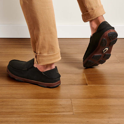 Olukai Moloa. Men's slip-on shoe in black waxed nubuck leather. Model wearing khaki pants from behind.